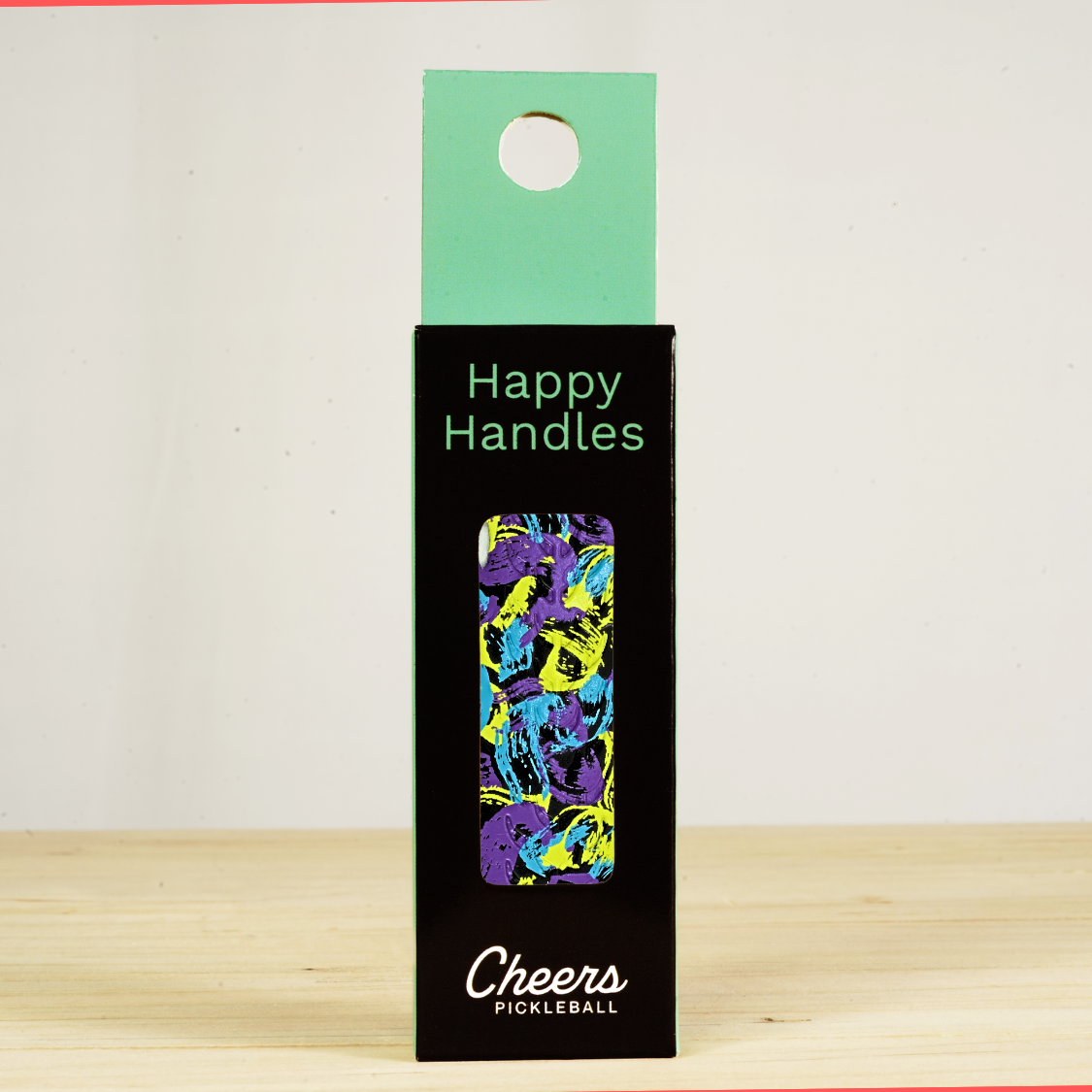 Happy Handles Overpriced Abstract Art Pickleball Grip in its packaging, featuring purple, yellow, and blue abstract paint design, visible through the product box window