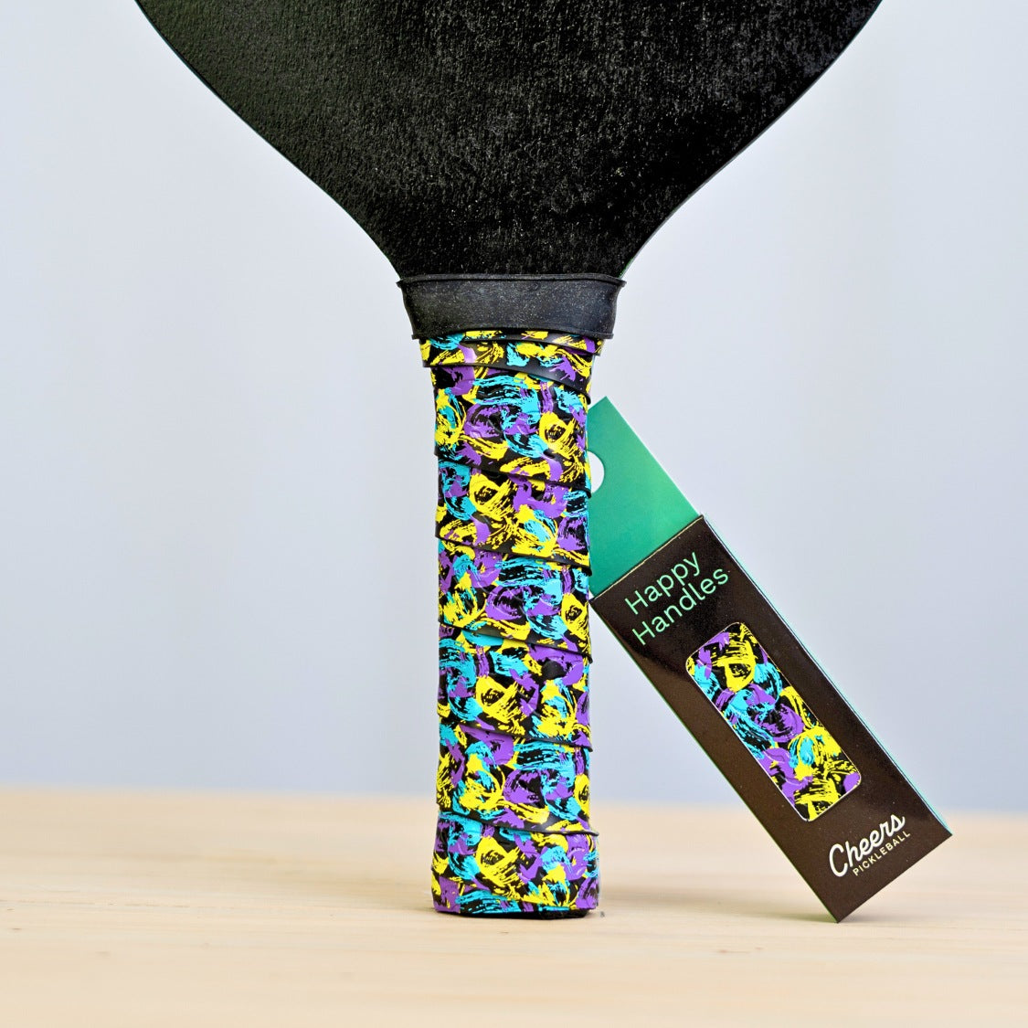 Happy Handles Overpriced Abstract Art Pickleball Grip with with purple, yellow, and blue abstract paint design displayed on a paddle
