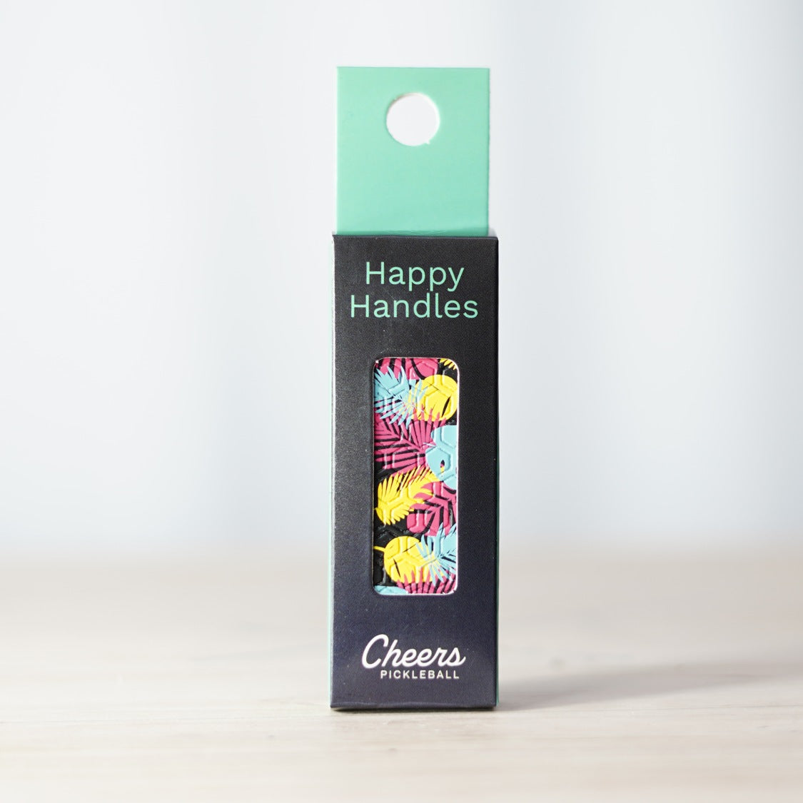 Happy Handles Rainforest Pickleball Grip in its packaging, featuring yellow, pink, and blue tropical leafs, visible through the product box window